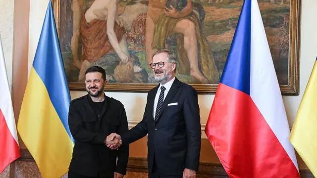 Петр Фіала у темному костюмі з краваткою і Володимир Зеленський у чорному стоять в Празі, вітаються за руку в приміщенні, де видно велику картину на фоні та великі українські й чеські прапори