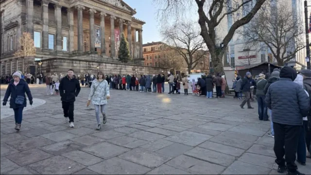 Una fila de decenas de personas frente a un edificio con columnas en el exterior. Se encuentran de pie sobre grandes losas de piedra, y la fila pasa junto a árboles sin hojas. Van vestidos para el invierno.