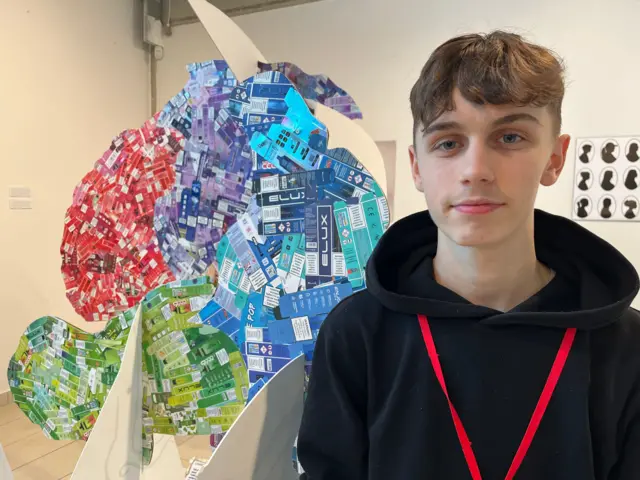 Un adolescente que viste una sudadera con capucha negra y un cordón rojo está parado frente a una escultura colorida hecha con envases de cigarrillos electrónicos vacíos.