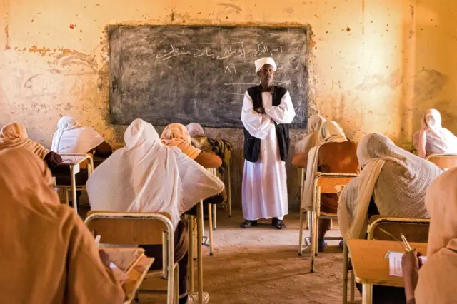 A teacher stands at the front of a classroom while invigilating an examination.