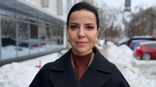 Oksana Dmitrieva se encuentra en una calle nevada de Kiyv. Lleva un abrigo negro y un jersey naranja/marrón.