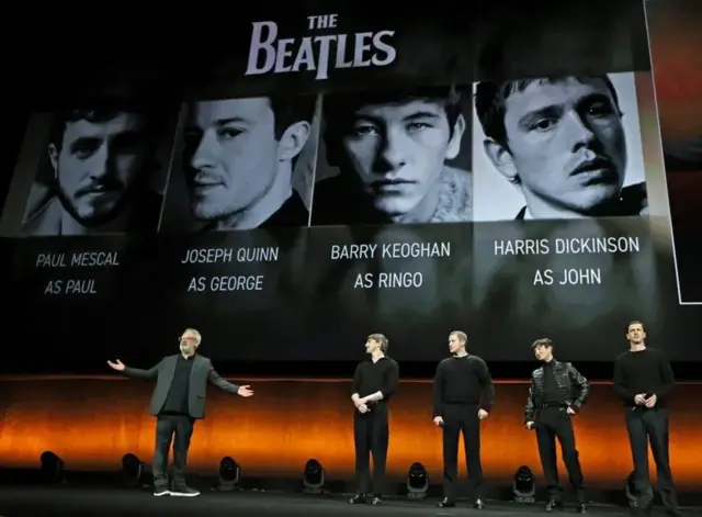 На сцене стоят режиссер и актеры, а на заднем плане - их фотографии