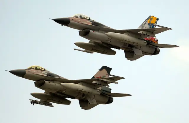 Dos cazabombarderos F-16 vuelan en formación cerrada, uno encima de otro en dirección hacia la izquierda. Ambos están pintados de colores de camuflaje, pero el de arriba tiene pintada la cola con los colores amarillo, azul y rojo, característicos de la bandera de Venezuela.