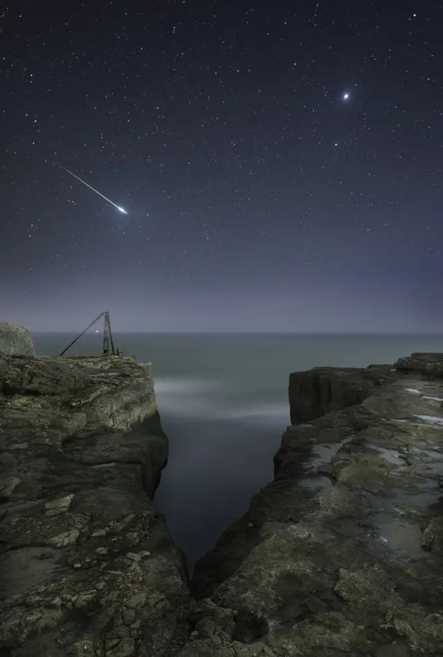 A shooting star flashes across the sky over the craggy landscape