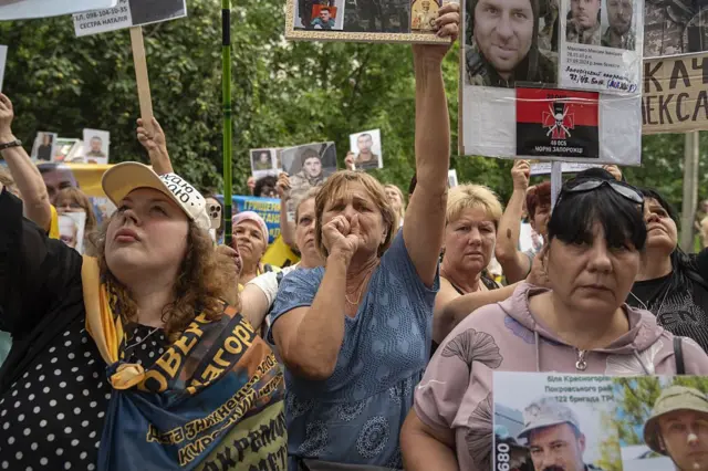 Жінки стоять щільною групою, багато з них тримають у руках плакати з великими кольоровими фотографіями чоловіків у військовій формі. На плакатах є імена, дати та підписи, деякі – з військовою символікою та прапорами. У центрі – старша жінка, яка пронизливо дивиться у камеру, притискаючи до губ стиснутий кулак