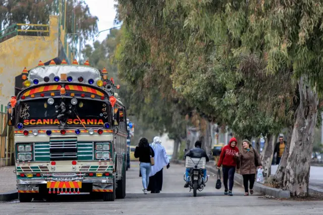 A la izquierda de una carretera, un autobús urbano lleno de farolillos y señas de colores. En la misma vía van caminando por el centro dos mujeres, una de ellas con velo en la cabeza, después una moto y otras dos mujers más. Al lado derecho de la vía hay una acera llena de árboles. 