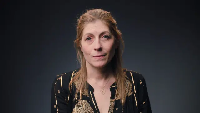 امرأة ذات شعر طويل بني فاتح مائل للحمرة تنظر إلى الأمام مباشرة. ترتدي بلوزة سوداء بخيوط ذهبية. وتضع حول عنقها نجمة داود داخل قلب.