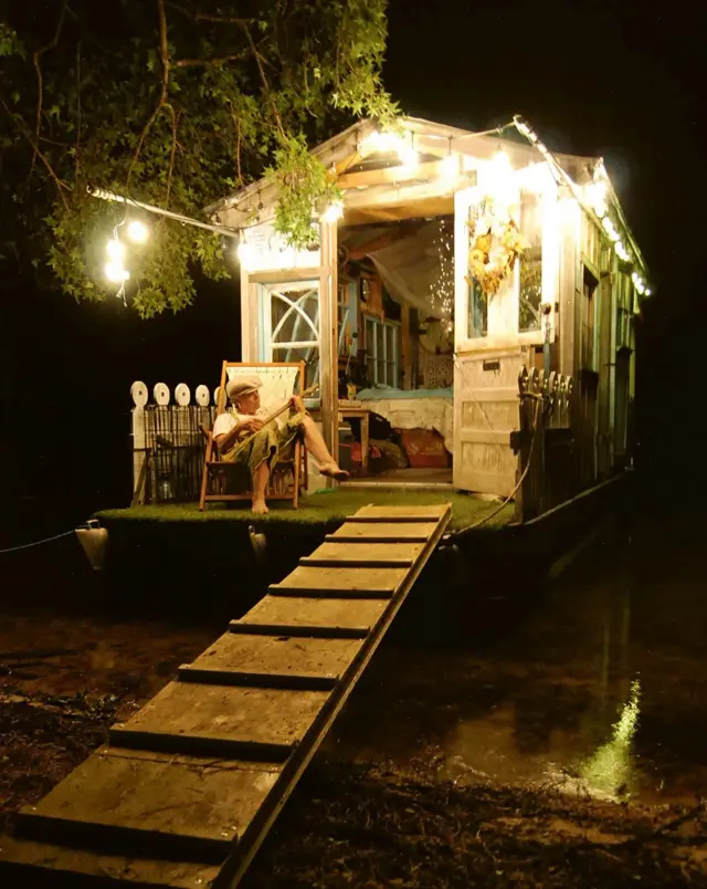 Foto mostra cabana em rio à noite iluminada por luzinhas e com uma mulher tocando banjo na varanda