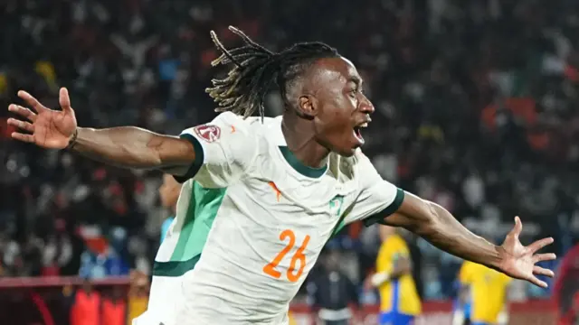 Yan Diomande, vêtu d'une chemise blanche ivoirienne avec un numéro 26 orange sur la poitrine, écarte les bras et ouvre la bouche en guise de fête, ses cheveux coulant derrière lui. Une foule bondée dans un stade de football est floue en arrière-plan