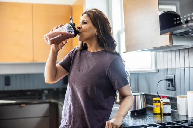 Una mujer mayor consume un batido de proteína en su cocina. 