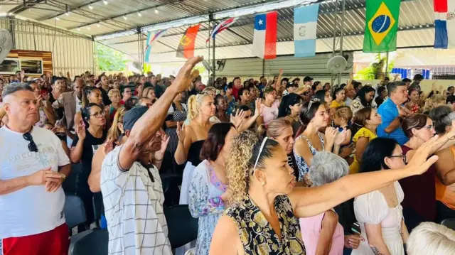 Pessoas com braços erguidos em igreja, em ambiente iluminado e com bandeiras de vários países, inclusive o Brasil