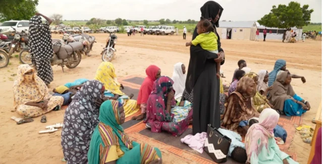 سيدات سودانيات يجلسن على الأرض في مخيم للنزوح ويحيط بها عدد سيارات الأمم المتحدة وخيام للنازحين.