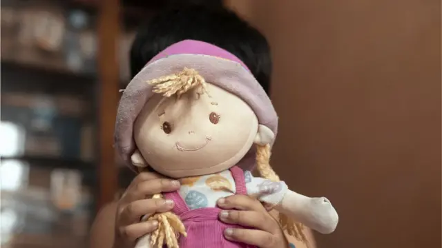 गुड़िया के पीछे चेहरा छिपाता बच्चा