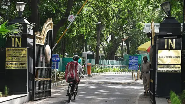 A uniformed guard stands at the gate of the Delhi Gymkhana Club as a cyclist zooms into the campus surrounded by trees. Plates on the gates and the wall mention "Members only", "Rights of admission reserved", "No thorough fare", "Trespassers will be prosecuted", etc. 