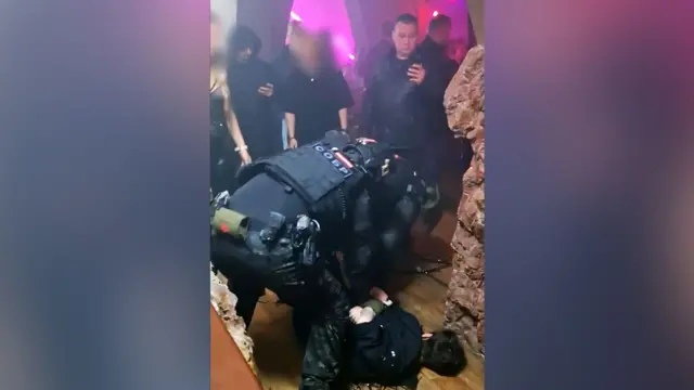 Rus kolluk kuvvetlerinden bir görevli, partinin yapıldığı mekânda yerde yatan ve elleri arkasında bağlı olan bir kişinin üzerine eğilmiş durumda, diğer konuklar ise olanları izliyor. Gece kulübünde pembe neon ışıklar yanıyor.