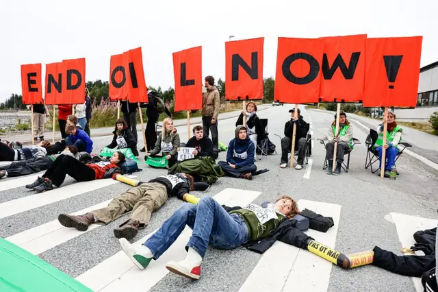 Un grupo de jóvenes protesta contra la explotación petrolífera en Noruega. Algunos están tumbados sobre el asfalto, otros sentados en sillas sostienen carteles con letras que unidas forman el mensaje en inglés "end oil now". 