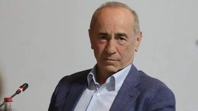 Koçaryan'ın katıldığı bir etkinlikte çekilen fotoğrafı. Üzerinde lacivert ceket ve açık renk bir gömlek var. Kıravat takmıyor.