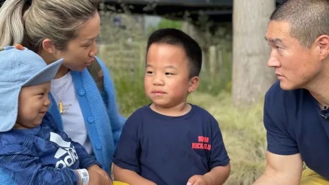 Oliver Chu, sorrindo, está no colo da mãe, Jingru. Ele usa boné e camiseta azuis e calças amarelas brilhantes. Jingru tem cabelo loiro preso em um rabo de cavalo e veste camiseta branca com cardigan azul. Com o outro braço, ela abraça o filho mais velho, Skyler, que veste camiseta azul com letras vermelhas. O pai, Ricky, está sentado ao lado de Skyler, usando camiseta azul e calças amarelas, com cabelo bem curto.