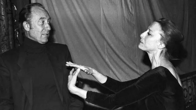 Черно-белая фотография: Щедрин в черной водолазке и пиджаке стоит в стороне, Плисецкая в одежде для репетиции балета практикует движения рук.