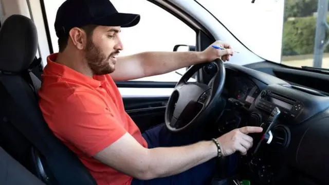 homem sentadosaque betwaybanco do motorista, com camiseta vermelha, mexesaque betwaycelular posicionado no painel do carro