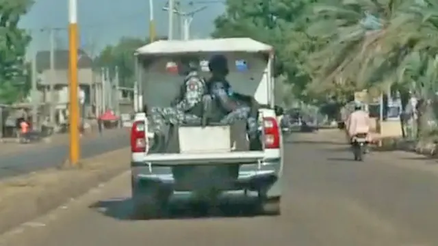 Un camion 4x4 de la police avec deux agents à l'arrière, servant d'escorte de sécurité, roule sur une route goudronnée bordée d'arbres dans le nord-ouest du Nigeria.
