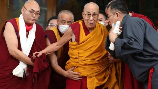 Tibetan spiritual leader Dalai Lama as dem dey assist am wen e arrive for an event for 2023, e wear yellow robe while im men wear black and wine robe.
