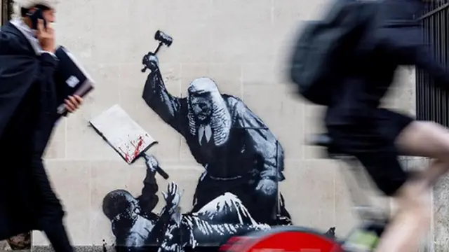 Картина Бэнкси на стене Высокого суда Лондона: судья в парике избивает молотком протестующего. На первом плане виден проходящий мимо судья в парике