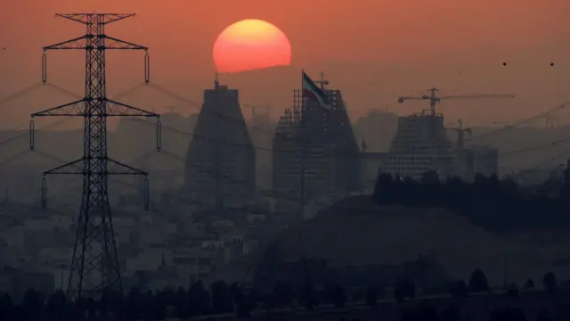 غروب خورشید در پشت ساختمان های بزرگ در حال ساخت غرب تهران در غروب، نشان دهنده رشد سریع املاک و مستغلات پایتخت ایران و خطوط انتقال نیروی برق است.