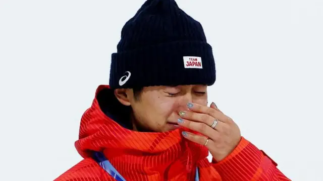 金メダル獲得に感極まった様子の戸塚。「Team JAPAN」と白地に赤で描かれた黒いニット帽と、オレンジ色のウェアにメダルの青いリボンが見える。目元を押さえている左手に、中指と小指に銀と金の指輪をしているほか、中指の爪に金色のラメ、ほかの指の爪に銀色のラメのネイルをしている 