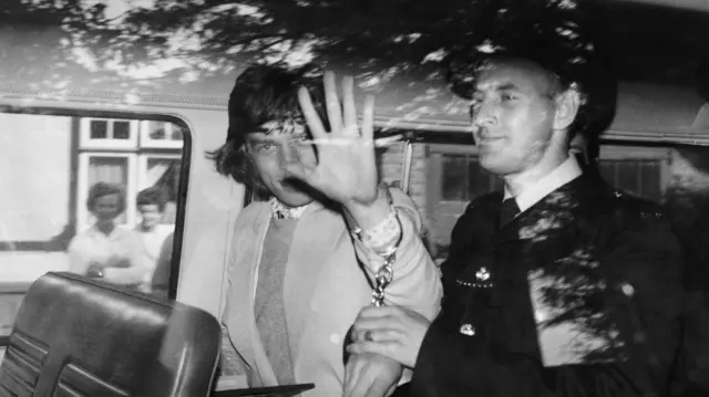 Police taking away Mick Jagger after Jagger was found guilty of the illegal possession of pep pills in 1967. Mick Jagger is in handcuffs and putting a palm up to the camera