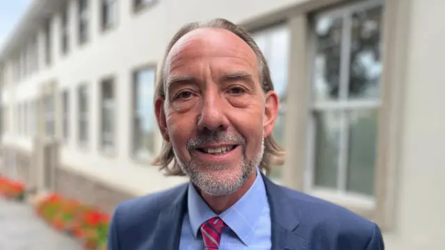 Diputado Steve Falla: Un hombre de pelo largo y castaño, con barba gris e incipiente, vestido con traje azul, camisa azul y corbata a rayas rojas y azules.  