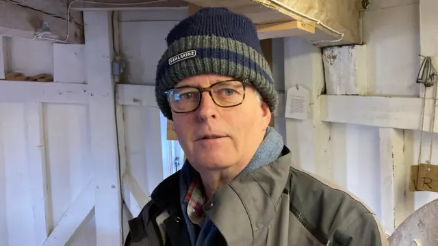 Kelvin Law, un hombre que se encuentra dentro del Molino Wicken, mira directamente a la cámara. Lleva un sombrero azul y verde, un impermeable verde y gafas protectoras.