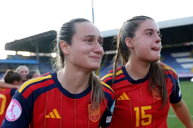 Kızıl renkli İspanyol formalarıyla iki kadın futbolcu gözyaşlarıyla sevinç yaşıyor. Uzakta bir yere doğru bakıyor gibiler.