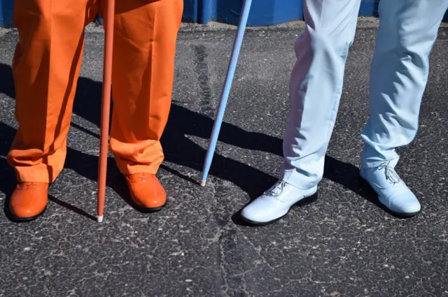 One man wears orange trousers, shoes and holds an orange cane, with the man next to him wearing all pale blue