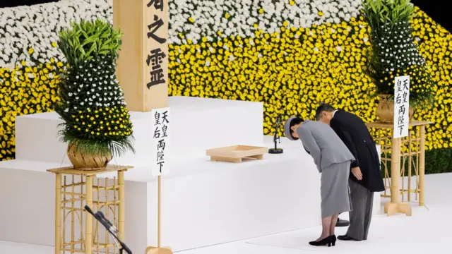 Императорская чета (Масако в сером костюме, серой шляпе и черных туфлях; Нарухито в темно-серых брюках и черном сюртуке) кланяются перед мемориалом, сделанным из дерева. На стене — белые и желтые цветы.