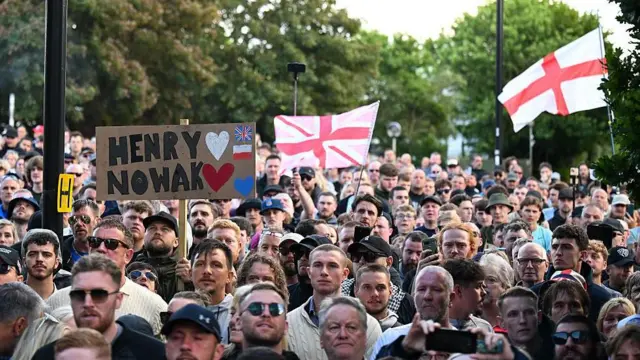 Grupa ludzi protestujących, trzymających transparenty z nazwiskiem Henry'ego Nowaka i machających flagą Anglii.
