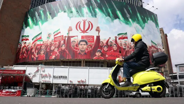 德黑蘭瓦利亞斯廣場一輛黃色摩托車駛過一座大樓上懸掛的巨型政治宣傳海報,海報中一群身穿紅色衣服的群眾舉起伊朗國旗與國徽(6/4/2026)