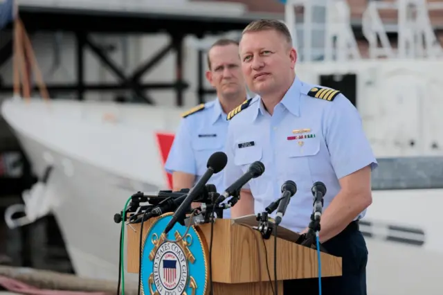Jason Neubauer falando no microfone em um púlpito ao lado de outro militar fardado