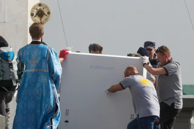 Отгрузка Владимирской и Донской икон Богоматери из собрания Государственной Третьяковской галереи у храма Христа Спасителя: несколько грузчиков переносят большую запакованную икону. Рядом священник, на заднем плане виден полицейский