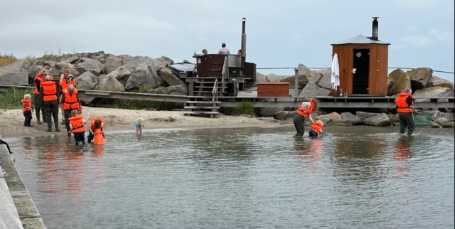 Varias personas, algunas con chalecos salvavidas naranjas, se adentran en aguas poco profundas con redes junto a una orilla rocosa, donde también se ven una sauna de madera y una pequeña construcción sobre una plataforma.