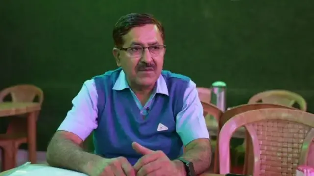 মোহাম্মদ আখলাকের ভাই জান মোহাম্মদ টেবিলে হাত রেখে একটি প্লাস্টিকের চেয়ারে বসে আছেন 