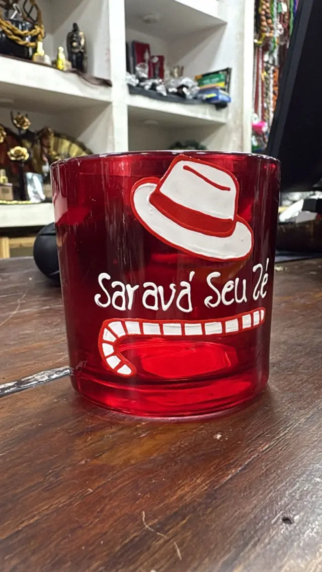 Copo transparente com a palavra “Saravá” impressa em vermelho, exposto em prateleira de loja de artigos religiosos. Ao fundo, outros objetos de culto afro-brasileiro, como imagens e velas coloridas.