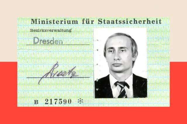 Thẻ căn cước Stasi màu xanh nhạt có ảnh đen trắng của ông Vladimir Putin ở bên phải. Ông mặc bộ vest tối màu và thắt cà vạt caro, tóc vàng và đang nhìn về phía xa, hướng ra ngoài khung hình.