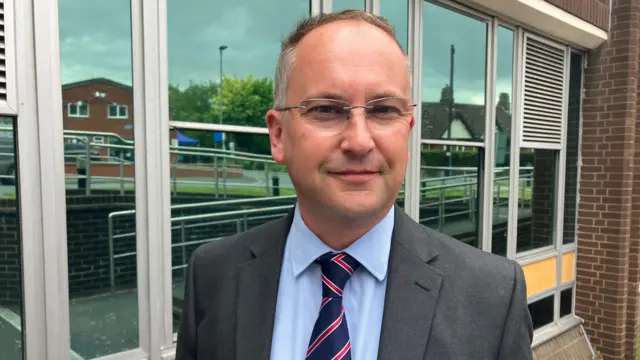 Un hombre que vestía un traje gris con una camisa azul y una corbata roja y azul, estaba detrás de un moderno edificio con fachada de cristal.