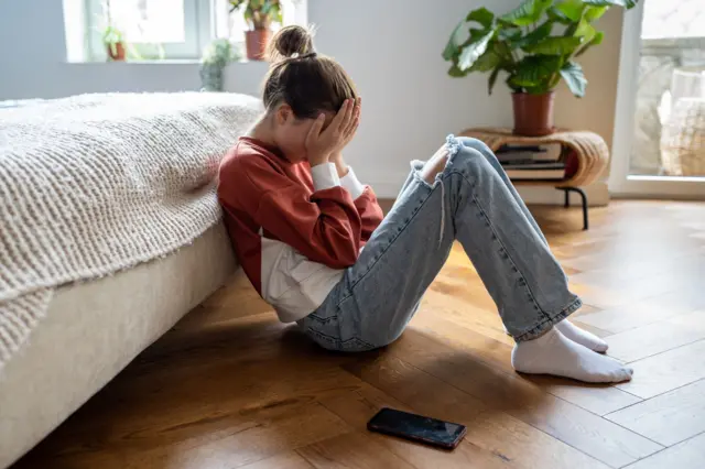 Jovem sentada no chão do quarto, com as mãos no rosto e o telefone celular jogado ao lado.