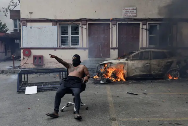 Demonstrant sa maskom preko lica i bez majice sedi ispred auta koji gori