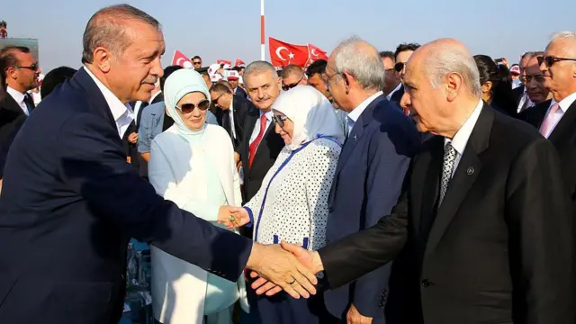 Cumhurbaşkanı Erdoğan ve MHP Genel Başkanı Devlet Bahçeli, darbe girişimine karşı 7 Ağustos 2016'da İstanbul Yenikapı'da düzenlenen mitingde el sıkışıyor. Arkada Emine Erdoğan, Binali Yıldırım, Kemal Kılıçdaroğlu ve Semiha Yıldırım görülüyor. Arkada daha ileride ise Türk bayrakları taşıyan bir kabalalık bulunuyor.