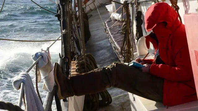 Giulia Baccosi escreve seu diário no convés do navio, em alto mar. Ela veste um casaco vermelho com capuz.