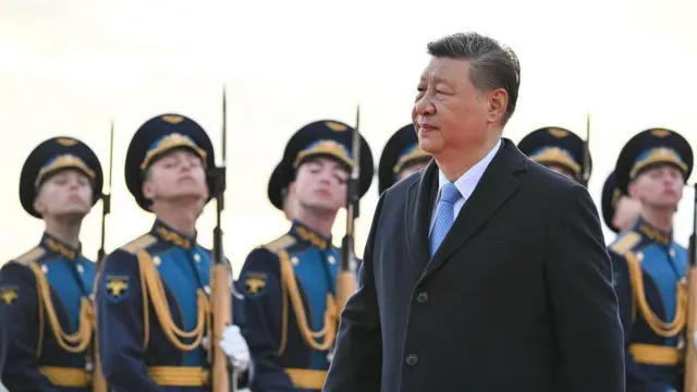 Xi Jinping wearing a black coat and light blue tie, walking in front of a row of Russian soldiers carrying bayonets
