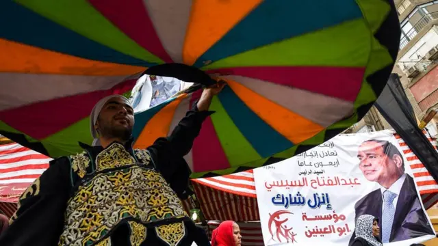 A whirling dervish dances past a poster of incumbent President Abdel Fattah al-Sisi wuth a caption reading in Arabic 'go and participate', outside a polling station in the capital Cairo's western Giza district on 28 March 2018.